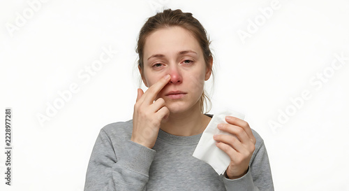 A single young adult woman experiencing common cold symptoms, holding a tissue in one hand and gently rubbing the side of her nose with the other