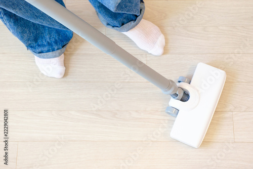 Top view of a female legs in jeans and white socks, with a vacuum cleaner on white laminate flooring.