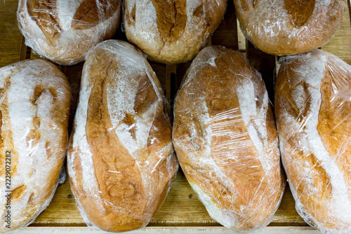 Several freshly baked bread loaves wrapped in transparent plastic wrap displayed on a wooden surface. The breads have a golden-brown crust with flour dusting and distinct scoring patterns. The arrange