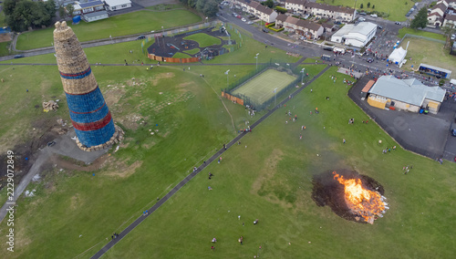 Aerial view of a large bonfire structure and a smaller burning fire in a park with a tennis court and residential houses in Larne, Northern Ireland, United Kingdom.