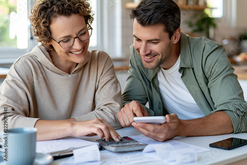 Couple smiles while managing household finances together. Woman taps calculator, man holds phone. Both look pleased, focused, and engaged. Scene suggests budgeting, planning, or savings