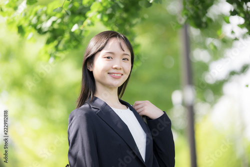 春の新緑が美しい屋外を笑顔で微笑む、新卒の若い日本人女性社員、大学生の就活や新入社員としてフレッシュなイメージのカメラ目線笑顔