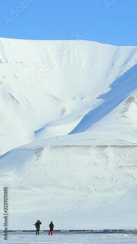 Two tiny silhouettes of people standing on a frozen shore against a massive snow-covered mountain under a clear blue sky in Svalbard archipelago Norway