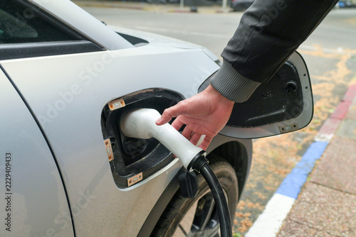 Close up of a hand plugging a white charging connector into the port of a silver electric car parked on a city street, with the charge port cover open and a black cable hanging.