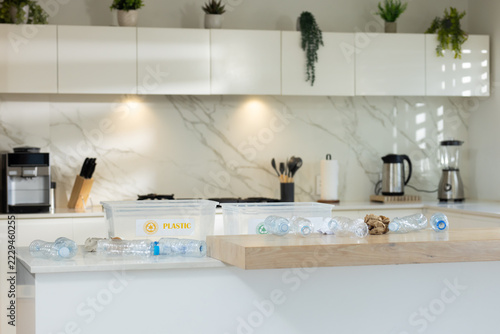 Clear plastic bottles are scattered on white kitchen island near recycling bins with PLASTIC label