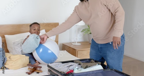 African father, youth son packing suitcase on bed, child handing backpack, father closing for trip