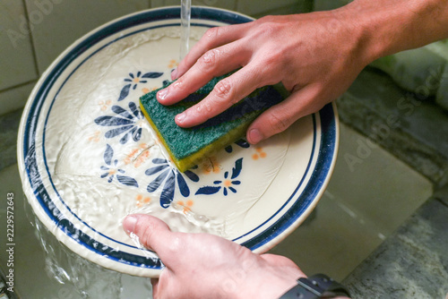 Close up of hands scrubbing a blue and white floral ceramic plate with a green and yellow sponge under running tap water in a kitchen sink.