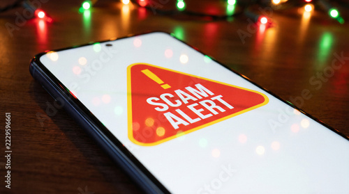 Smartphone displaying red scam alert warning on wooden table with colorful christmas lights highlighting holiday fraud risks.