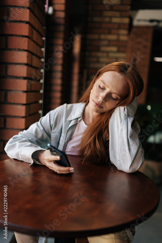 Exhausted young woman with long red hair rests head on hand at cafe table. Sleepy female with closed eyes holding smartphone, feeling burnout, boredom in cozy restaurant with warm lighting.