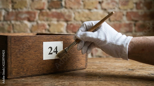 White gloved hand with paintbrush dusting antique wooden box with label number 24 on brick wall background