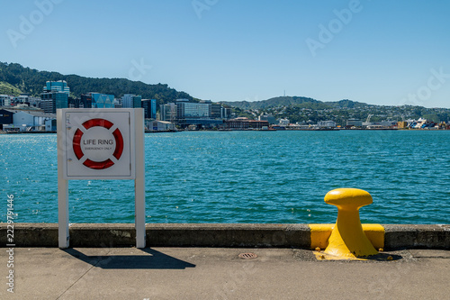Sunny summer day in Wellington, New Zealand showing the harbour, city and hills from the Taranaki Wharf. The yellow bollard and life ring are in foreground. No people