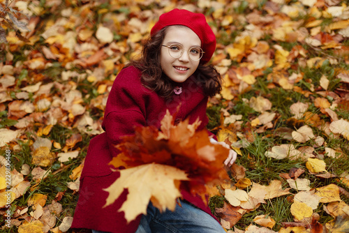 High-angle portrait of happy smiling young girl wearing red beret, glasses, and burgundy coat, sitting on ground covered with colorful yellow and orange fallen autumn leaves