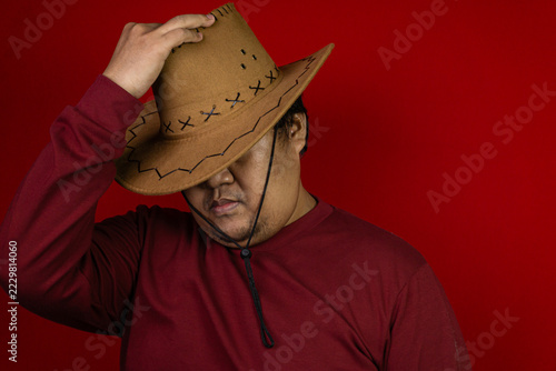 Obraz na plátně A stylish young Asian male pulling down the brim of his brown cowboy hat to partially hide his face, creating a mysterious and enigmatic atmosphere