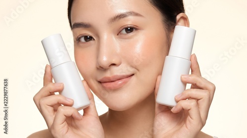 Beautiful Asian woman holding two blank white cosmetic bottle mockups near her face