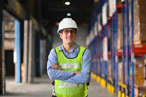 Smiling warehouse worker with safety helmet and reflective vest standing confidently in an industrial environment with shelves in a distribution center