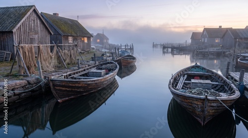 Wooden fishing boats moored gently alongside rustic weathered coastal cabins during a serene misty morning sunrise capturing peaceful Scandinavian maritime harbor scenery with calm reflective water su