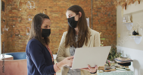 Caucasian female business owner and her coworker wearing face masks, using laptop and talking