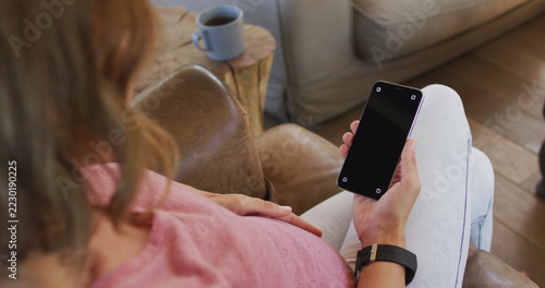 Happy caucasian pregnant woman sitting in armchair and using smartphone with copy space