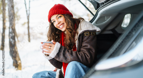 Woman in brightly colored hat and scarf sits on edge of car trunk, pouring hot beverage from thermos into mug. Traveler enjoys snowy forest from back of vehicle. Concept of winter day, relaxation.