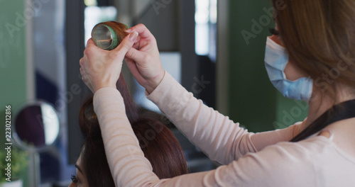 Causian female hairdresser in face mask styling hair of female client with curlers at salon