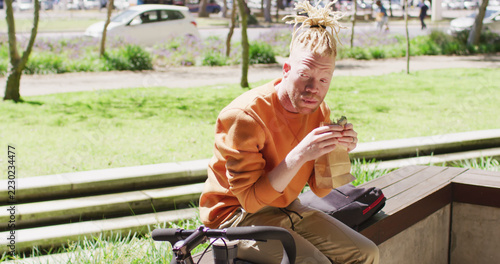 Albino african american man with dreadlocks sitting in park with bike eating sandwich