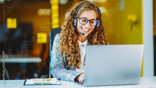 Young woman wearing glasses and headset works at computer in bright office. She is using headset with microphone while sitting at desk with laptop. Concept of support and technology.