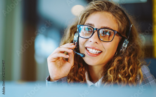 Young woman wearing glasses and headset works at computer in bright office. She is using headset with microphone while sitting at desk with laptop. Concept of support and technology.
