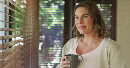 Happy caucasian pregnant woman standing at window with tea and touching belly