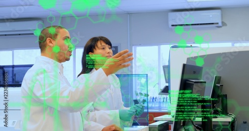 Gesturing tech showing monitors, partner watching at lab bench in lab coats green gloves and shield