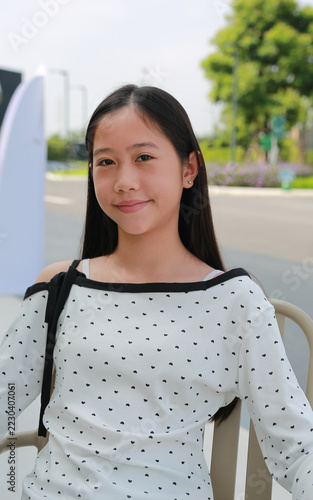 Smiling Asian preteen girl wearing a white off-shoulder top with heart patterns, sitting on a chair outdoors and looking at camera.