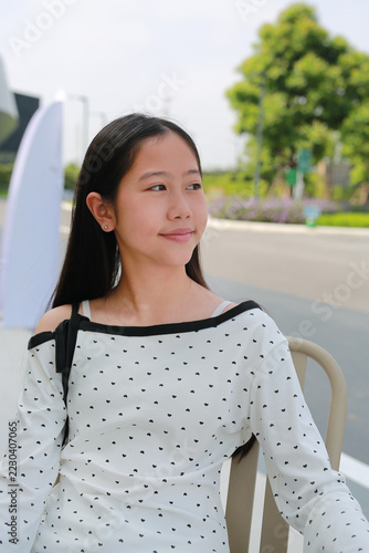 Smiling young Asian girl with long dark hair in white heart-patterned off-shoulder top sits on a chair outdoors, glancing to the side with a gentle smile on a sunny day.