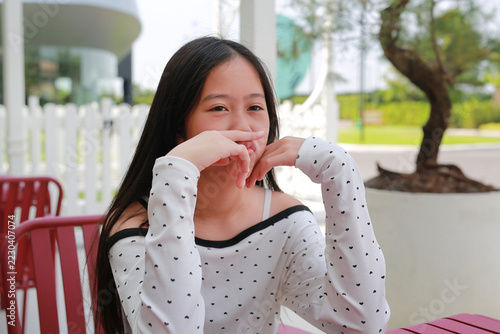 Asian preteen girl in white heart-patterned top, sitting at red outdoor cafe table and pressing her finger against her pursed lips in a thoughtful pose while looking camera.