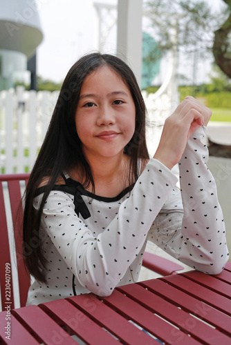 Smiling young girl with long dark hair in white heart-patterned top sits at a red outdoor table with her arms folded on it, looking at camera with a calm, confident smile.