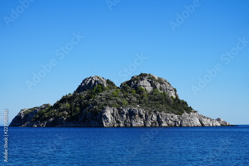 Rocky island with granite cliffs and scrubby vegetation surrounded by calm blue sea under clear sky near horizon view at water level, natural coastal landscape scene for travel and nature imagery