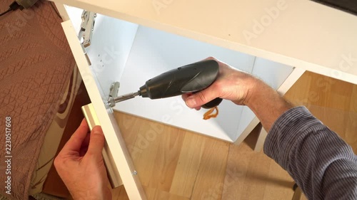 Installing metal hinge on white furniture panel with electric screwdriver. DIY furniture assembly or repair process.