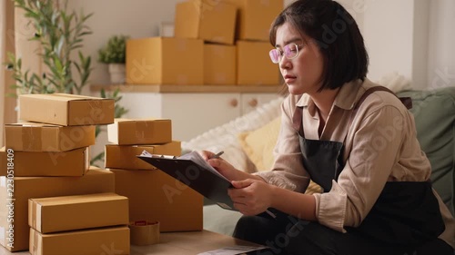 Asian female merchant wearing glasses holding clipboard, looking pensive while reviewing customer package delivery orders near parcel boxes.