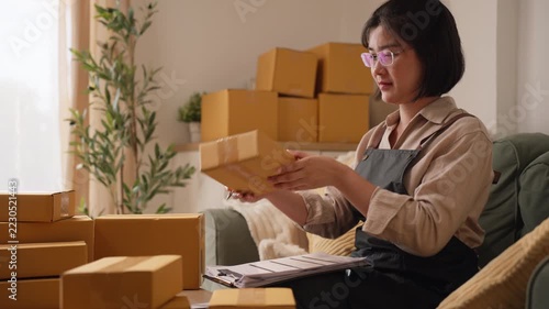 Asian female entrepreneur wearing glasses writing customer address details on paper while sitting near cardboard parcel boxes at home.