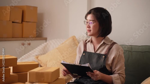 Asian female small business owner holding clipboard while stacking small cardboard boxes on desk, preparing for shipping dispatch.