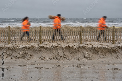 Dune fence on a beach with blurred crew in orange coats preparing for a noreaster, featuring wet gritty sand for coastal storm stories