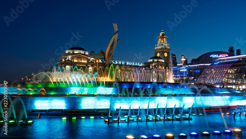 Illuminated fountain in front of Moscow railway station at night. Colorful water jets and historic station clock tower. Urban landmark and city travel. Public architecture and evening