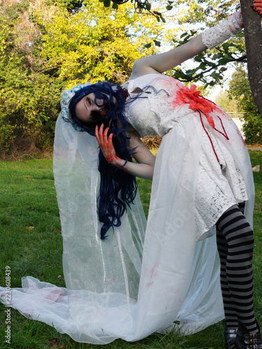 ghost bride bending backward holding tree in autumn park, bloody white wedding dress and long veil, halloween horror