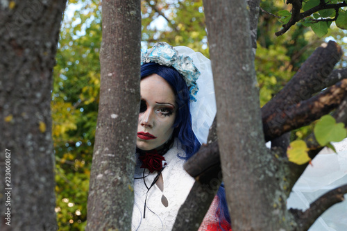 scary bride face peeking between tree trunks, blue flower crown and red rose choker, halloween horror portrait