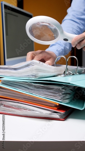 Business professional examining a thick stack of office documents with a magnifying glass, reviewing records for research, audit, verification, and financial analysis