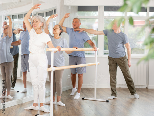 Diligent mature men and women practicing basic positions with trainer standing by ballet barre during workout session