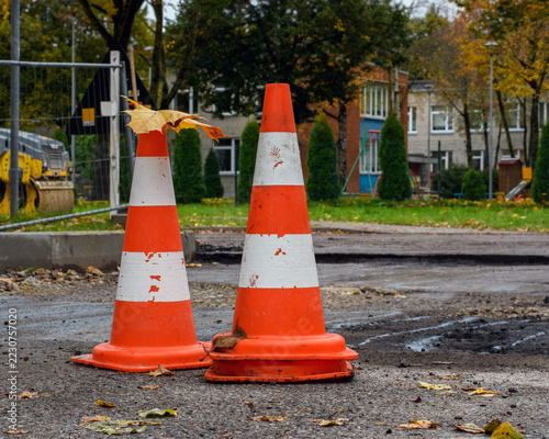 Two orange traffic cones with a fallen yellow maple leaf on top at a street repair site in a residential area during autumn.