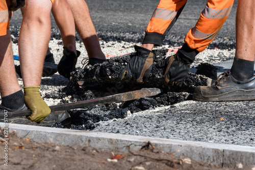 Close-up of road construction workers manually prying and lifting a layer of asphalt using a crowbar on a street repair site.