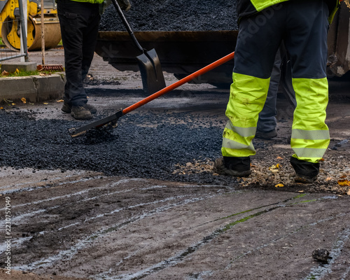 Close-up of a construction worker using a lute rake to level fresh hot asphalt mixture during street repair work.