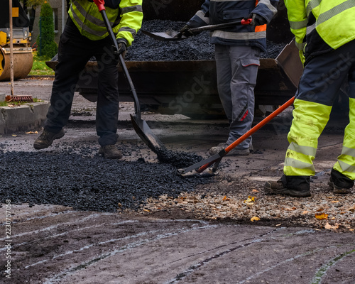 Close-up of road construction workers shoveling and spreading hot asphalt mixture with rakes from a loader bucket during street repair.