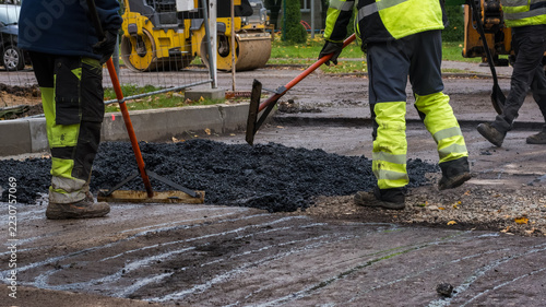 Road construction workers leveling freshly dumped hot asphalt mixture with rakes and lutes on a street repair site.
