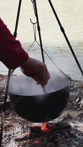 Cook removing cooked fish from a steaming tripod pot into a metal bowl outdoors, shot in vertical format.